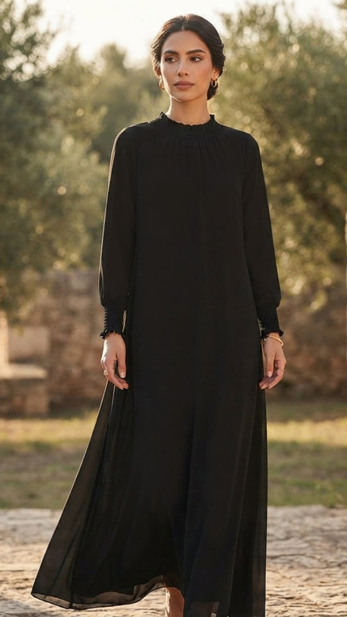 A woman wearing a long-sleeved black maxi dress stands outdoors in a sunlit garden setting with trees and a stone path in the background.