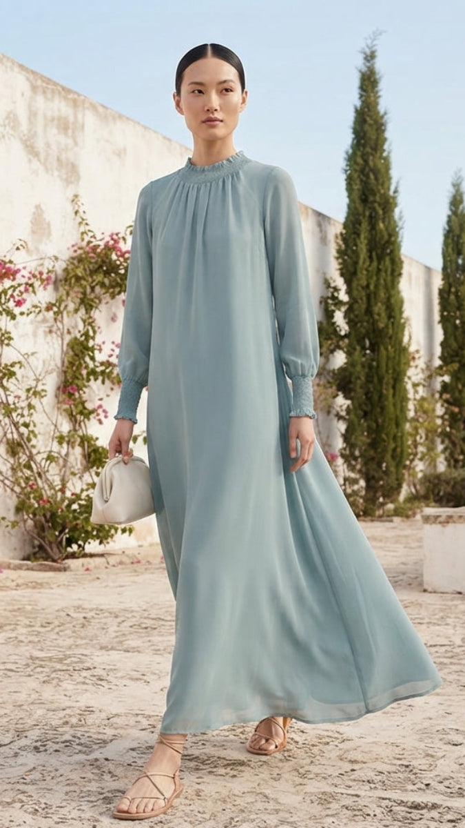 A woman modeling a long-sleeved, high-neck maxi dress in a dusty light blue-green color, standing outdoors against a white wall with climbing pink flowers and tall green cypress trees.