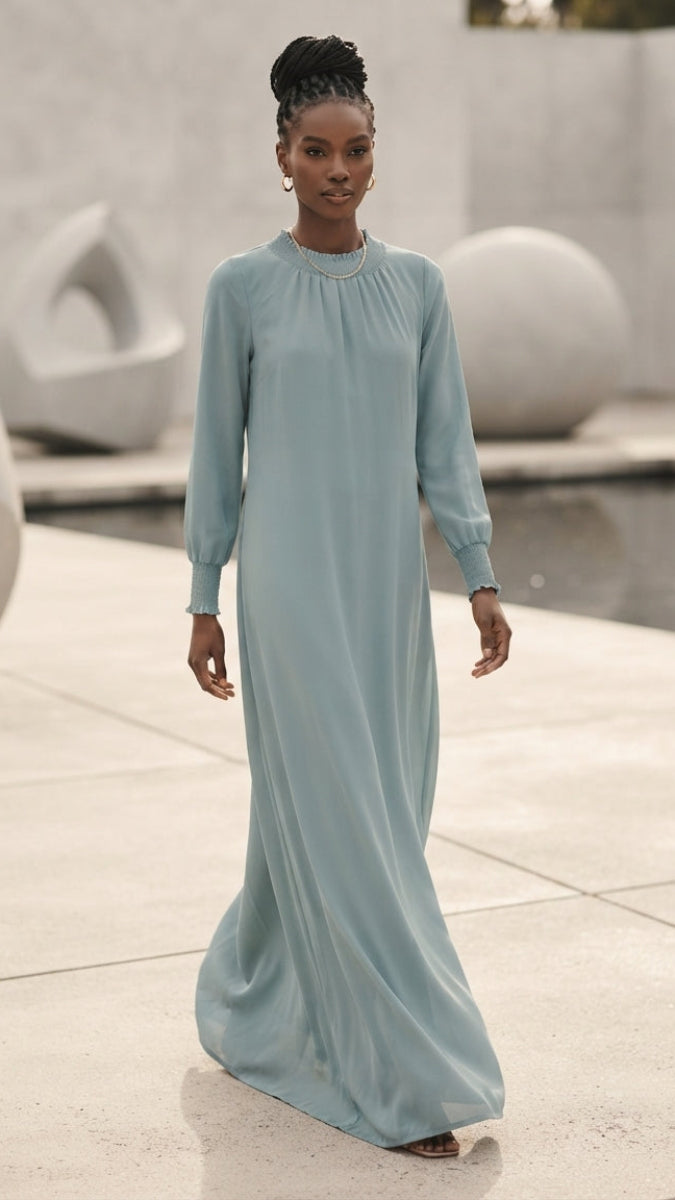 A woman wearing a long-sleeved, floor-length light blue dress walks outdoors on a paved area with modern sculptural planters and a low water feature in the background.