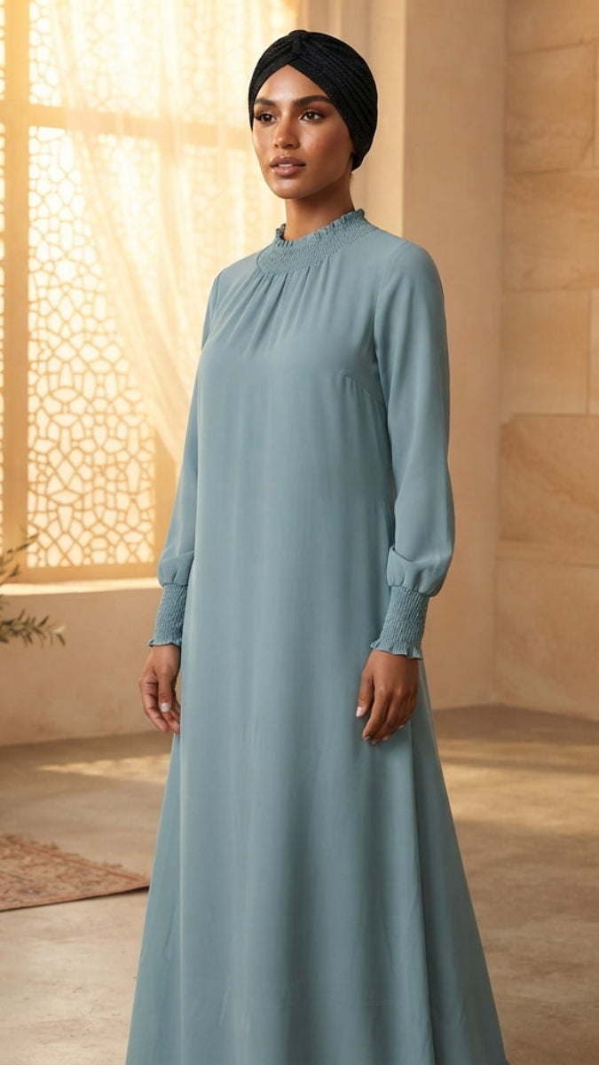 A woman wearing a long-sleeved, high-neck modest dress in dusty blue and a black hijab, standing in a warm, sunlit room with stone walls and potted plants.