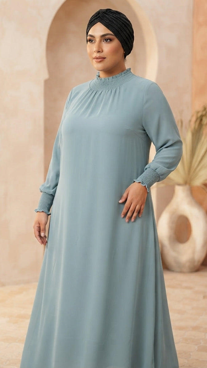 A woman wearing a long-sleeved, high-neck modest dress in dusty light blue, paired with a black headscarf, standing in a warm-toned architectural setting with arches.