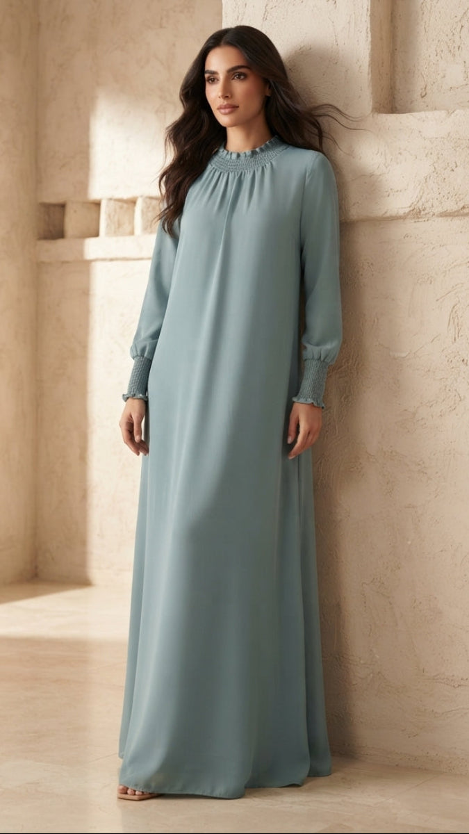 A woman wearing a long, flowing sage green maxi dress with long sleeves and a high gathered neckline, standing in a warm, sunlit architectural setting with textured beige walls.