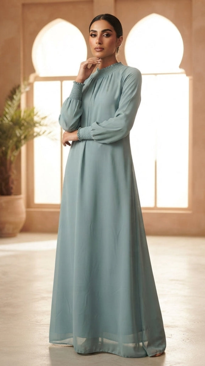 A woman modeling a long-sleeved, floor-length sage green dress in a warm, sunlit room with arched windows and potted plants.