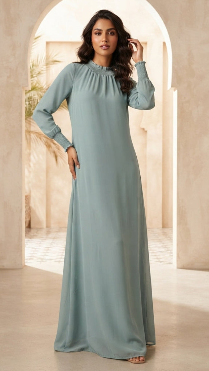 A woman modeling a long-sleeved, floor-length sage green dress in a warm, sunlit architectural setting with beige plaster walls and arches.