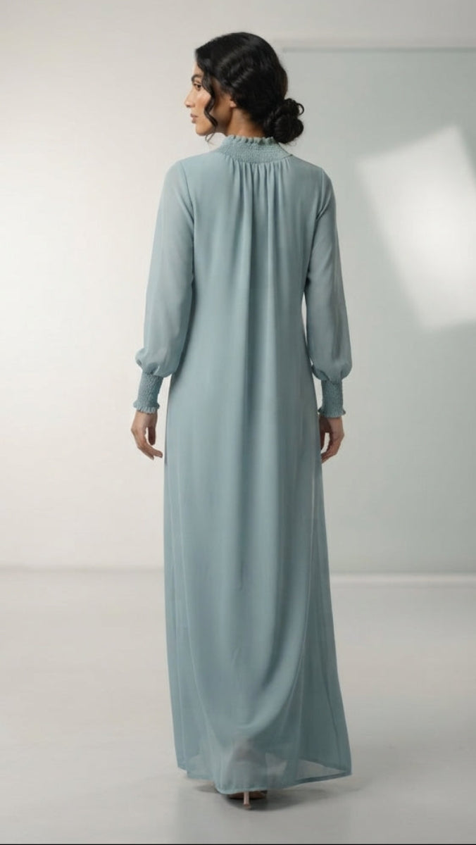 A woman stands with her back to the camera wearing a long, flowing light blue dress in a studio setting with softbox lights visible around the room.