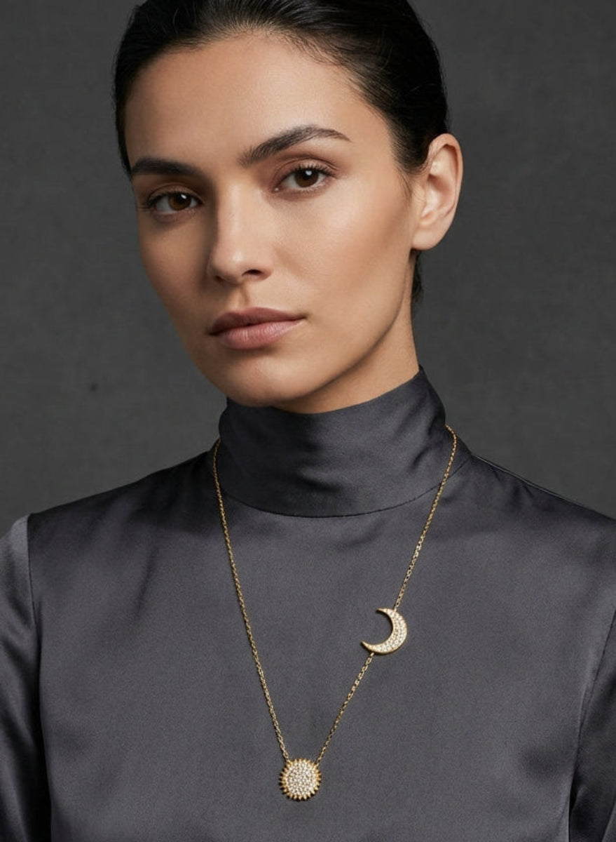 Woman wearing a dark gray blouse with a gold necklace featuring a moon pendant against a dark background
