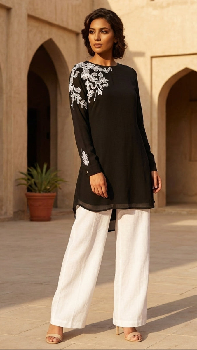 Woman wearing a black top with white embroidery and white pants in an outdoor setting with arches and plants.