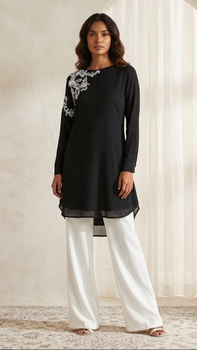 Woman wearing a black top with floral embroidery and white pants in a room with light-colored walls and a large window.