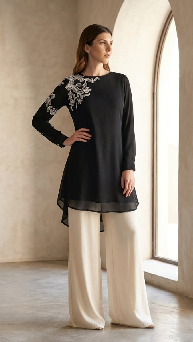 Woman wearing a black top with white embroidery and beige pants in a room with large windows.