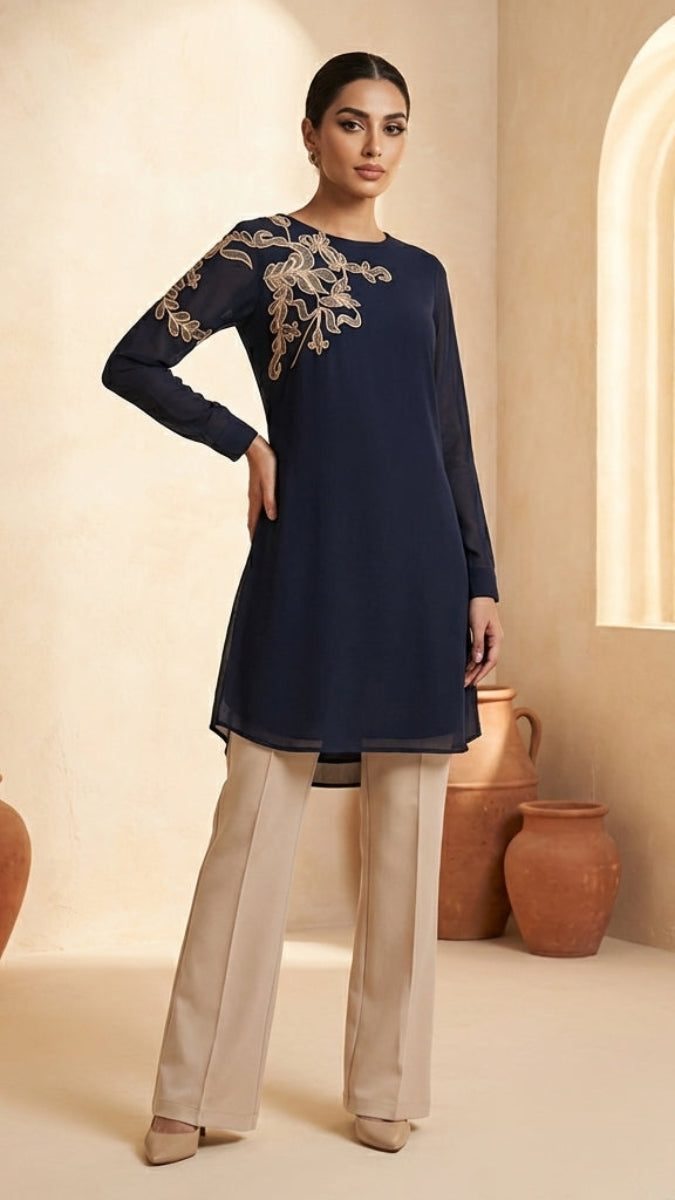 Woman wearing a navy blue embroidered top and beige pants in a warm-toned room.