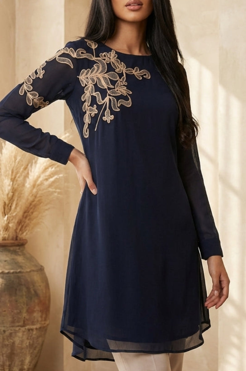 Woman wearing a navy blue dress with gold embroidery in a warm-toned interior setting.