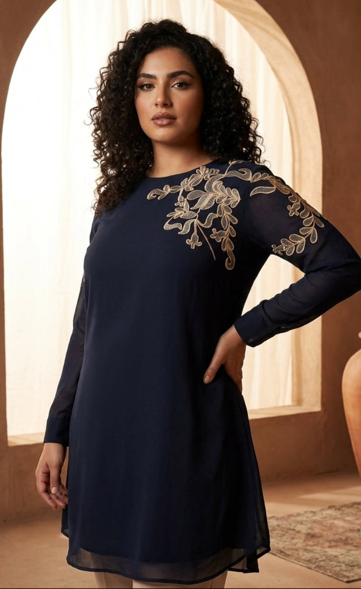 Woman wearing a navy dress with gold embroidery in a warm-toned room.