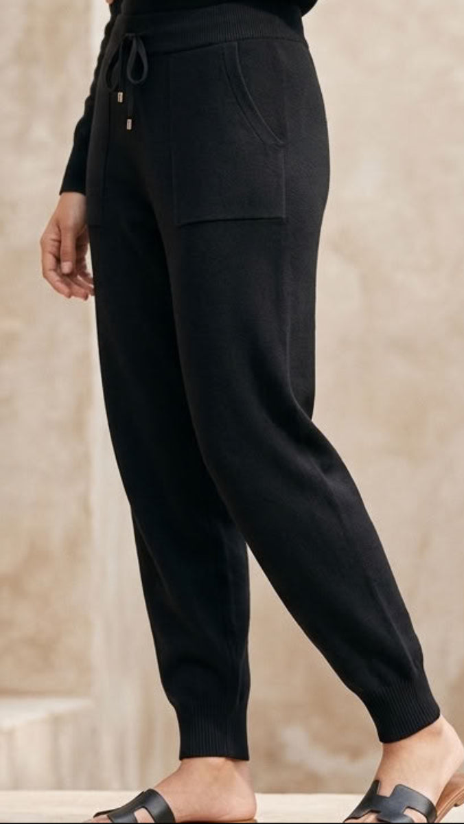 Black knit jogger pants with a drawstring waist and side pockets, shown on a model from the side against a neutral beige background.