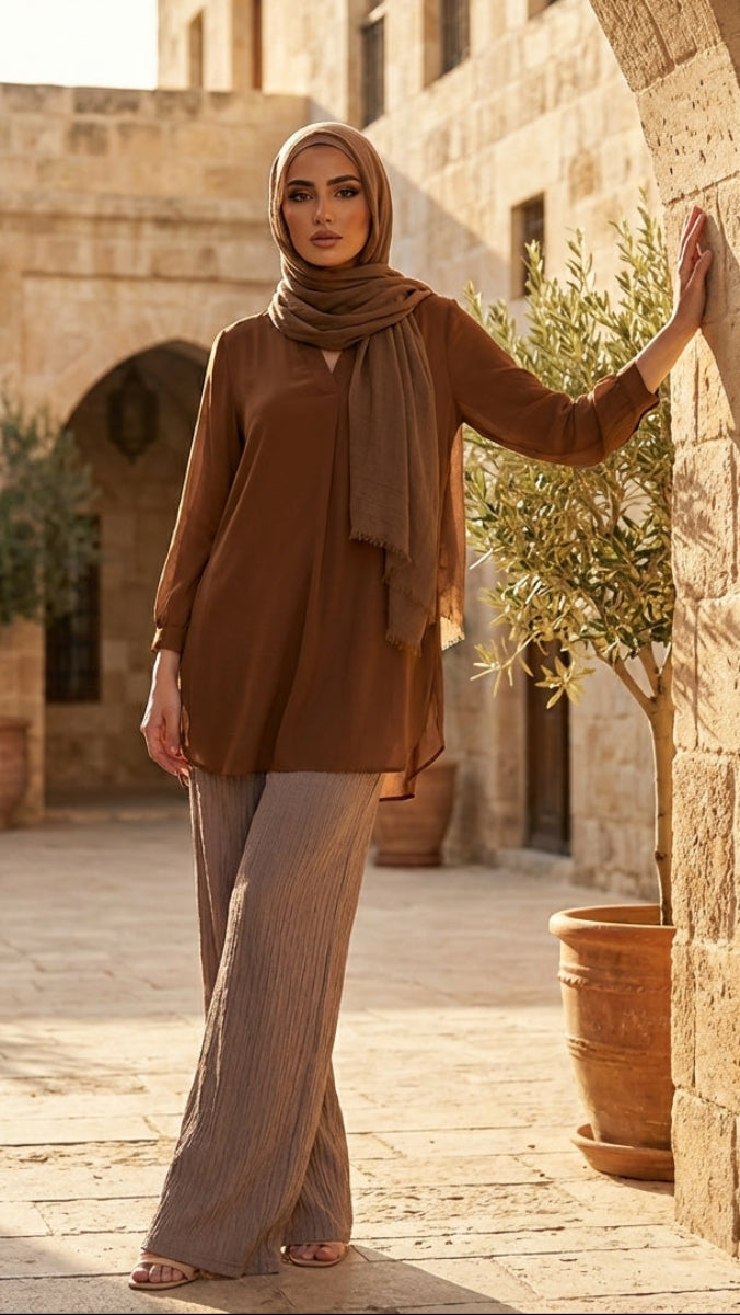 Woman in a brown hijab and outfit standing in a sunlit stone courtyard.