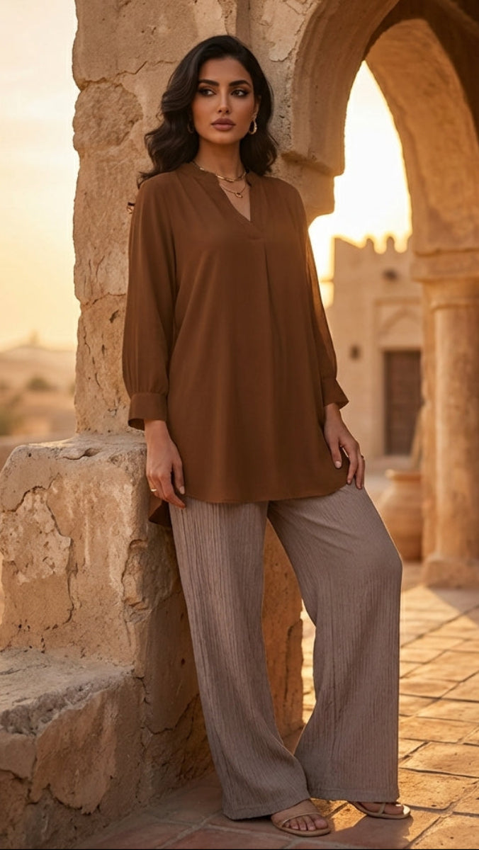 Woman in a brown top and beige pants standing in front of a stone archway with a warm glow.