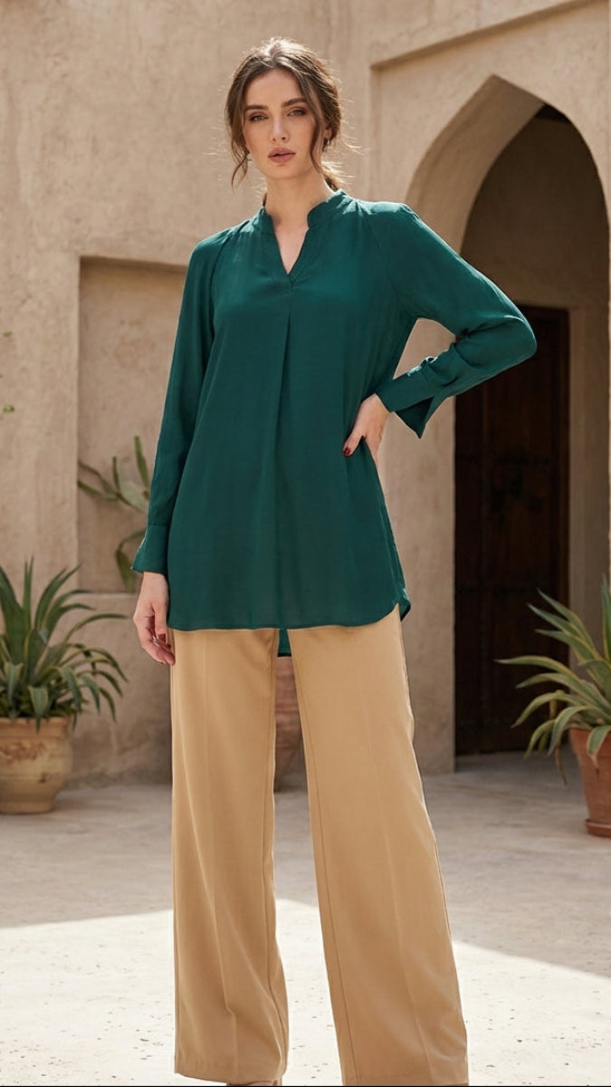 Woman wearing a green blouse and beige pants standing in front of a beige building with plants.