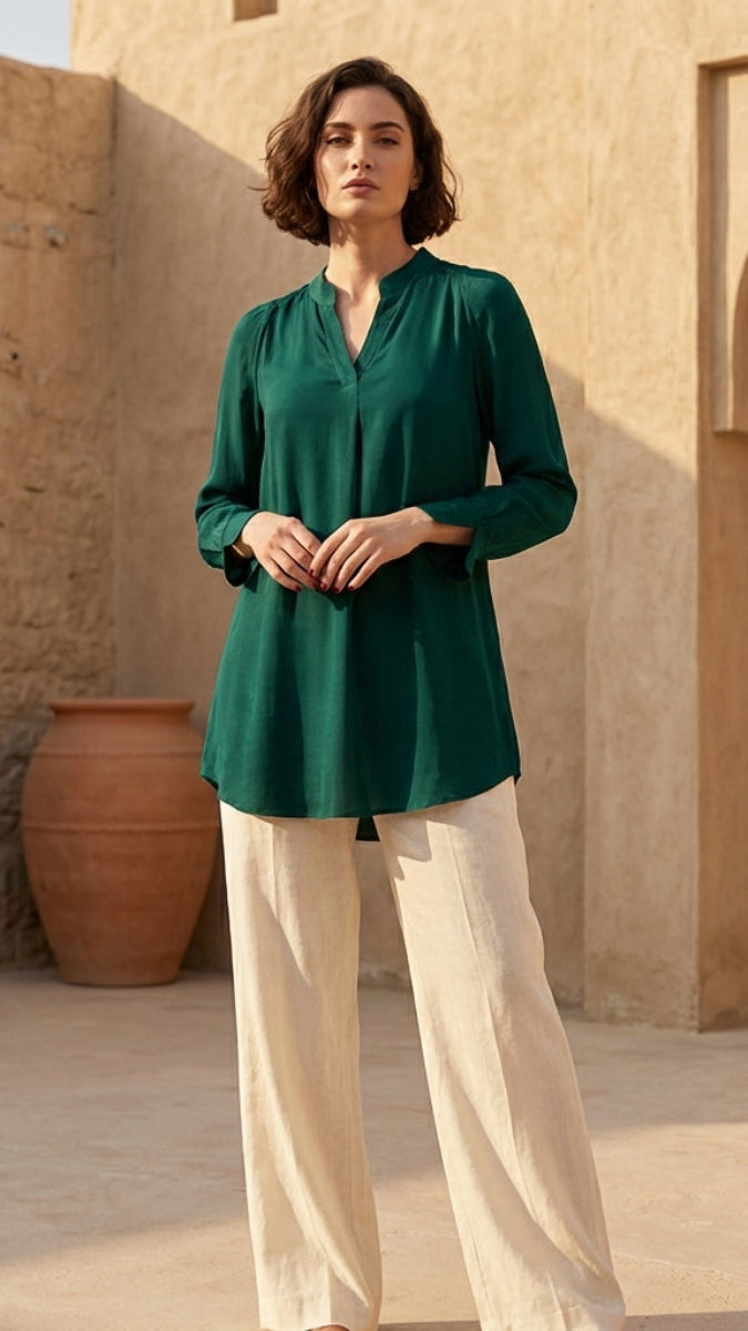 Woman wearing a green blouse and beige pants standing in front of a stone wall with an archway.