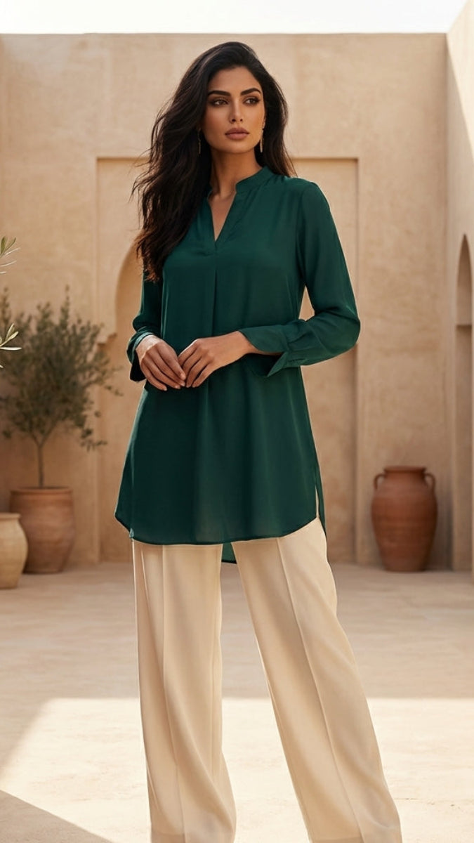 Woman in a green top and beige pants standing in a sunlit outdoor setting with architectural elements and plants.