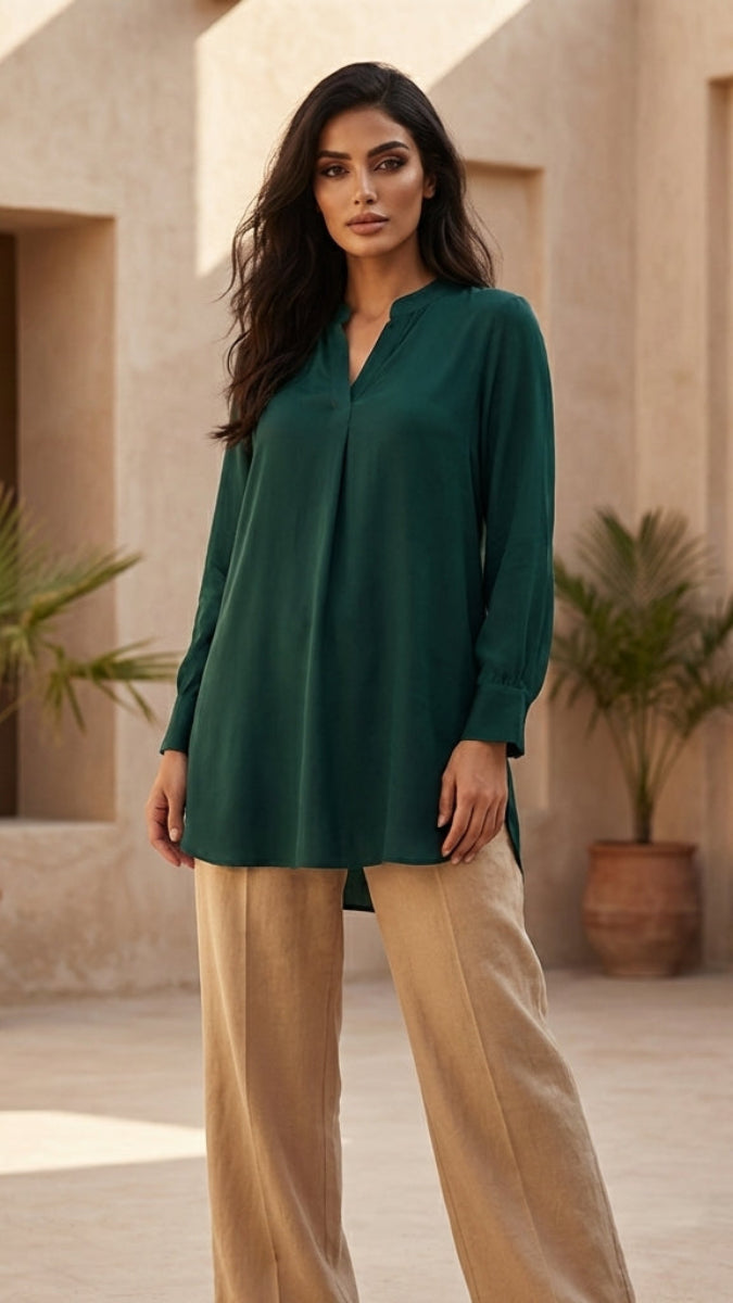 Woman wearing a green blouse and beige pants standing in a sunlit outdoor setting with architectural elements and plants.
