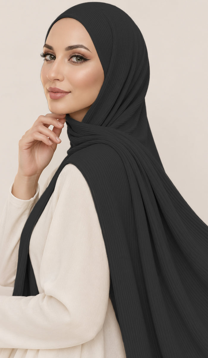 A woman wearing a black ribbed hijab and a cream-colored long-sleeve top, posing against a solid light beige background.