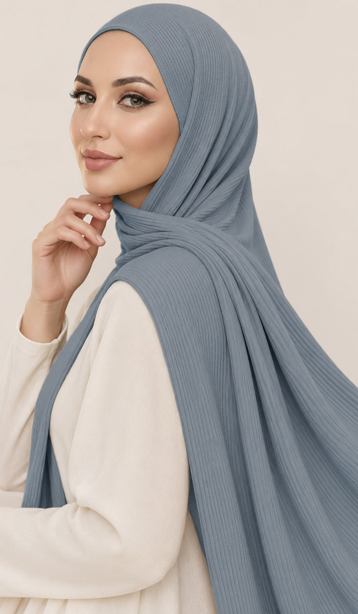 A woman wearing a slate blue-gray pleated hijab and a cream-colored long-sleeve top, posing against a solid light beige background.