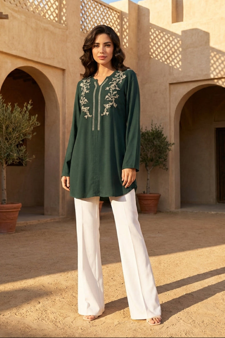 Woman in a green embroidered top and white pants standing in front of a traditional architectural background.