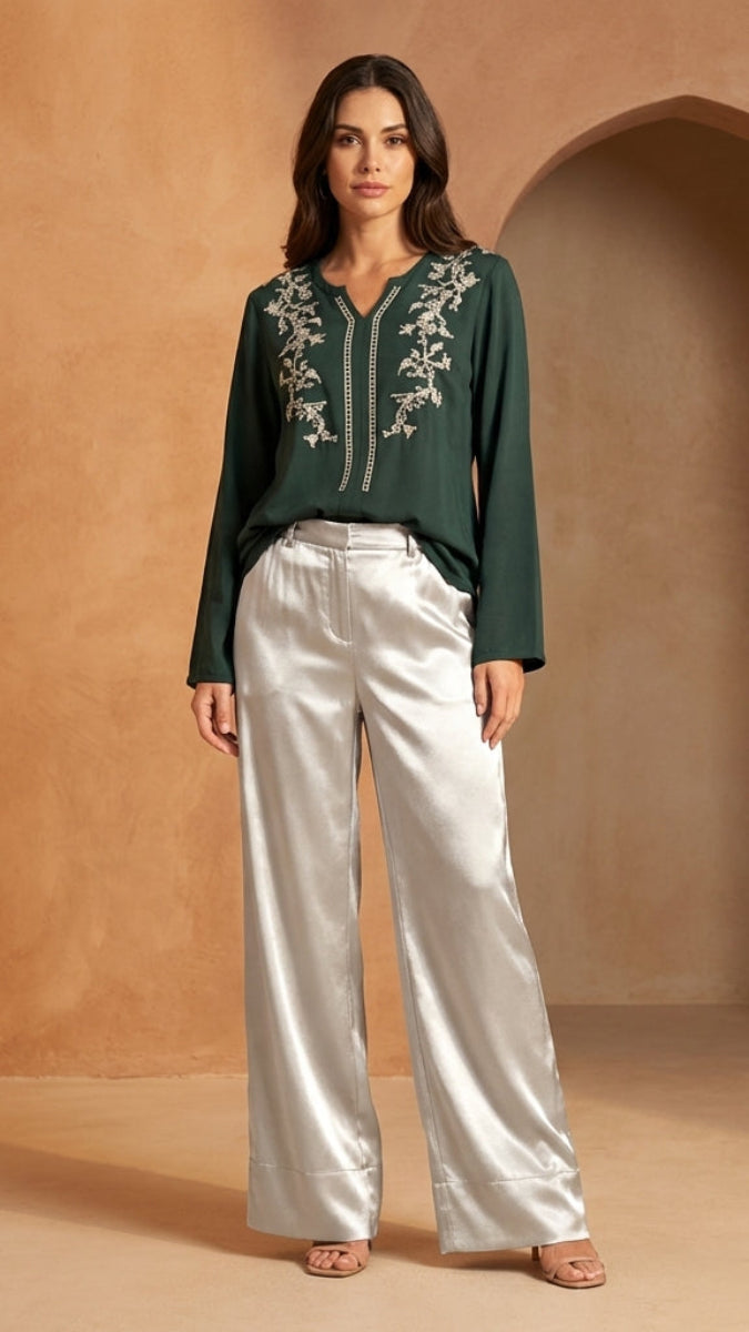 Woman wearing a green embroidered top and white pants standing in a warm-toned room with an archway.