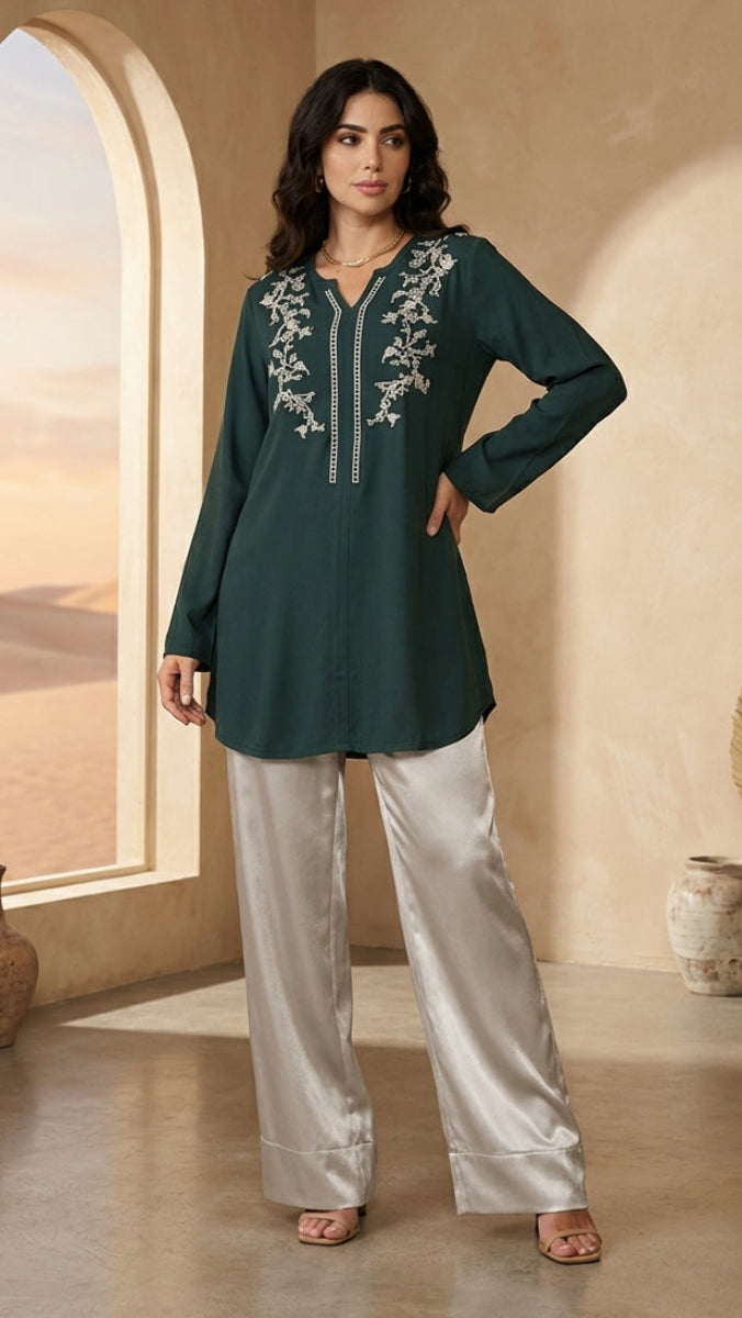Woman wearing a dark green embroidered top and light gray pants in a warm-toned room.