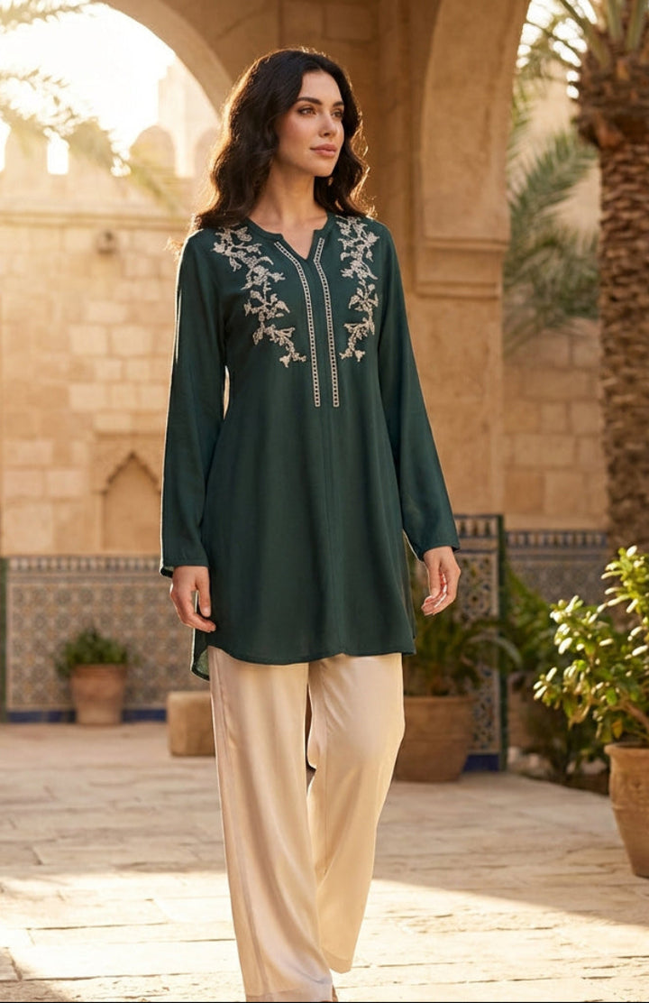 Woman in a green embroidered dress walking in a sunlit courtyard with palm trees and architectural elements.