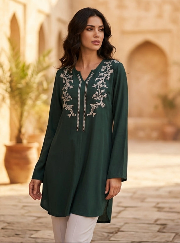 Woman in a green embroidered dress standing in a sunlit courtyard with arches and potted plants.