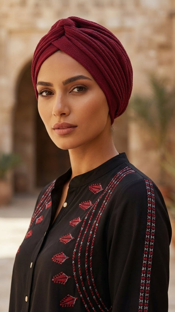 Woman wearing a maroon turban and black embroidered outfit in an outdoor setting with stone architecture and plants.