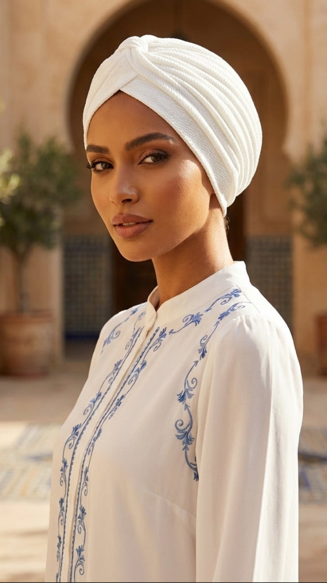 Woman wearing a white embroidered dress and headscarf in an outdoor setting with arches and plants.