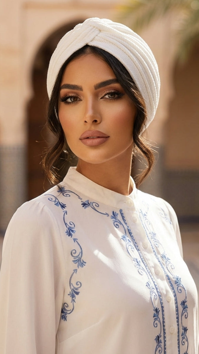 Woman wearing a white embroidered top and headscarf in an outdoor setting with architectural elements.