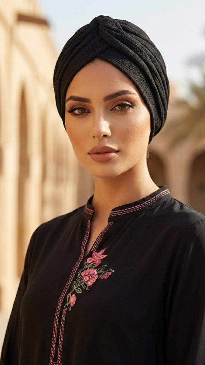 Woman wearing a black embroidered top and headscarf in an outdoor setting with palm trees.