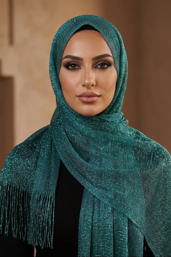 A woman wearing a shimmering teal green hijab with fringe detailing, styled over a black top against a warm, softly blurred architectural background.