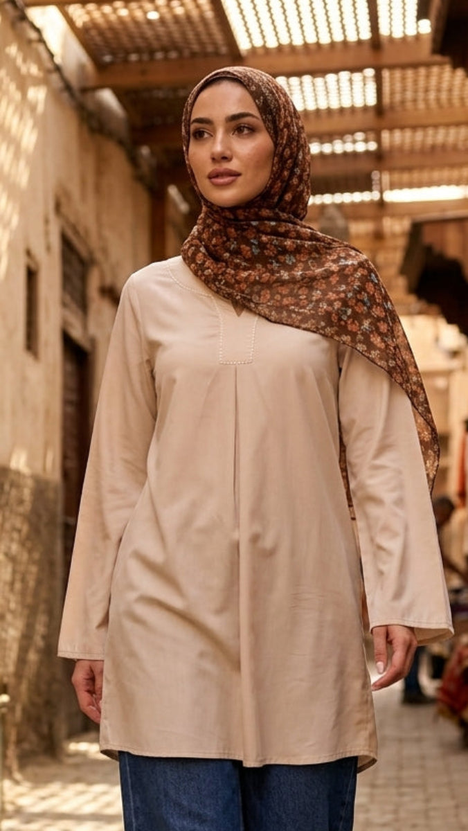 Woman in a beige coat and patterned hijab walking through a narrow street with traditional architecture.