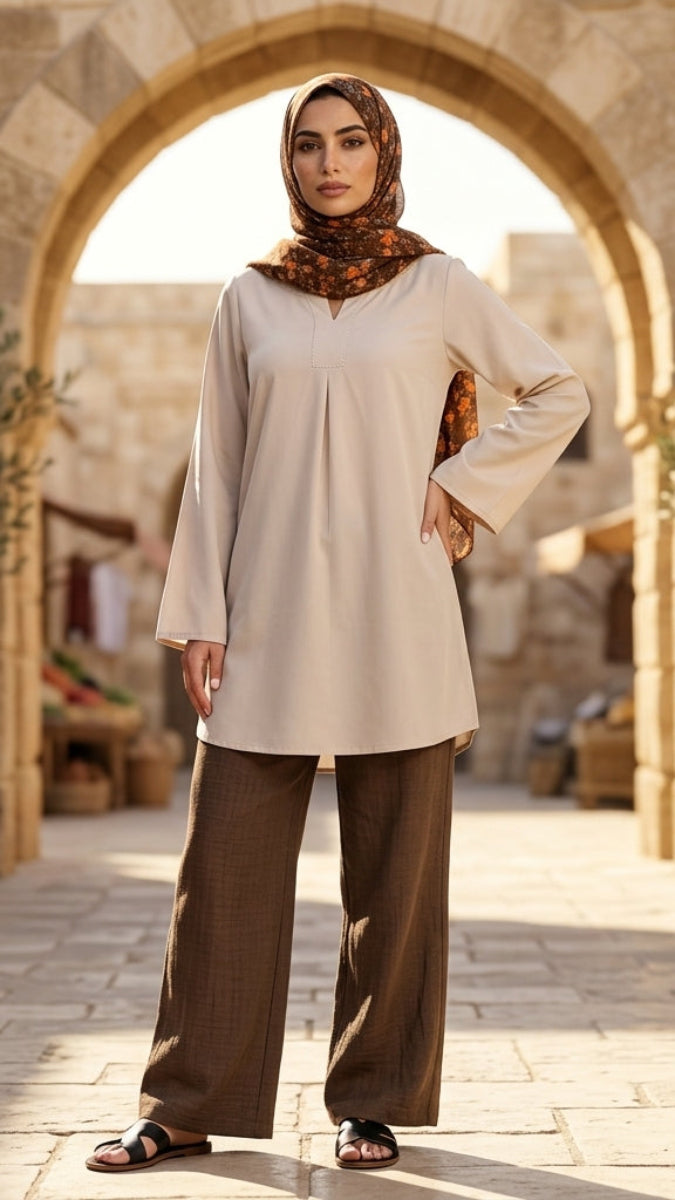 Woman in beige top and brown pants standing in a sunlit outdoor setting with stone archway and potted plants.
