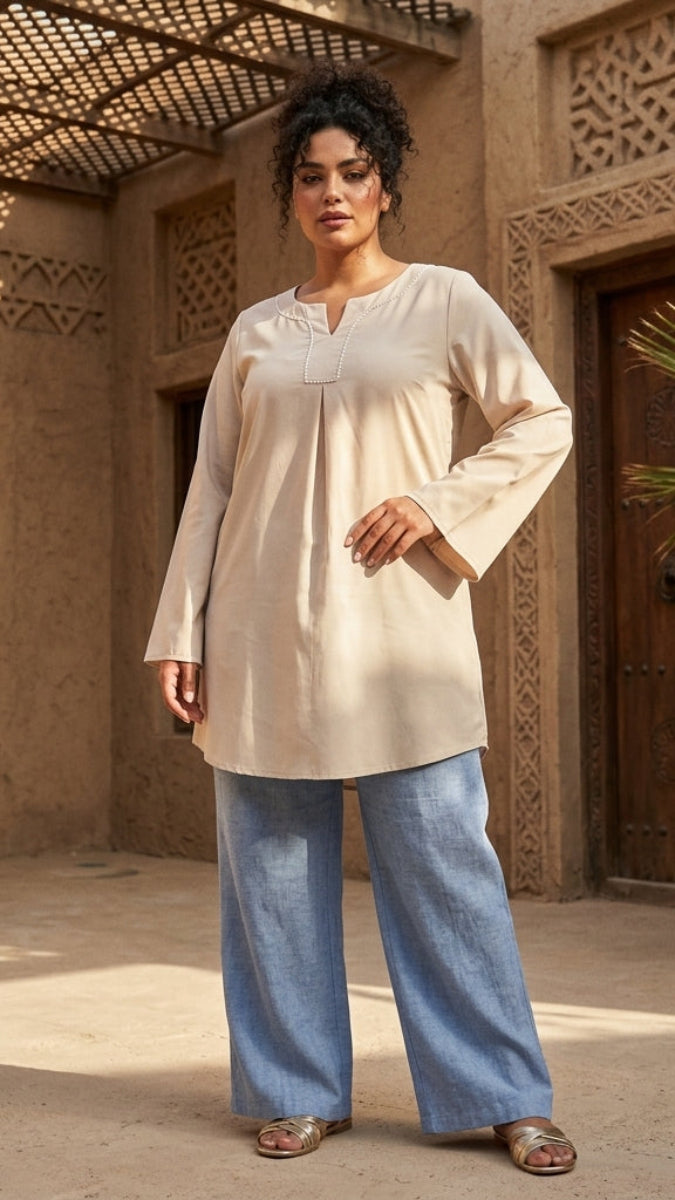 Woman wearing a beige top and blue pants standing in a sunlit courtyard with architectural elements.