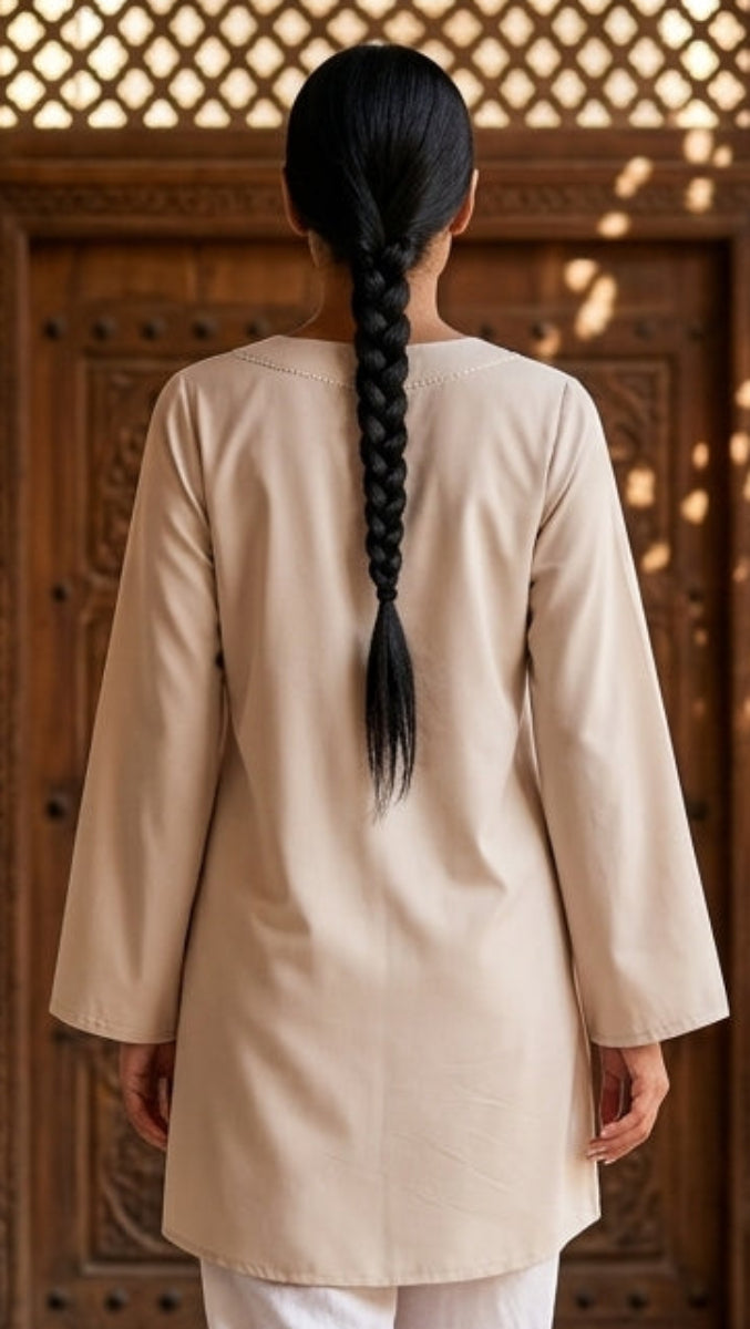 Person in a beige coat and white pants walking through a sunlit hallway with decorative tiles.