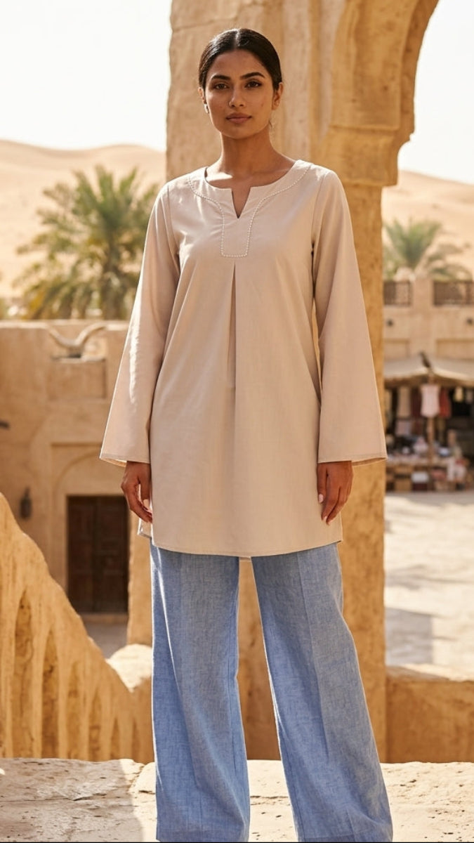 Woman in a beige top and blue pants standing in a desert setting with arched stone buildings.