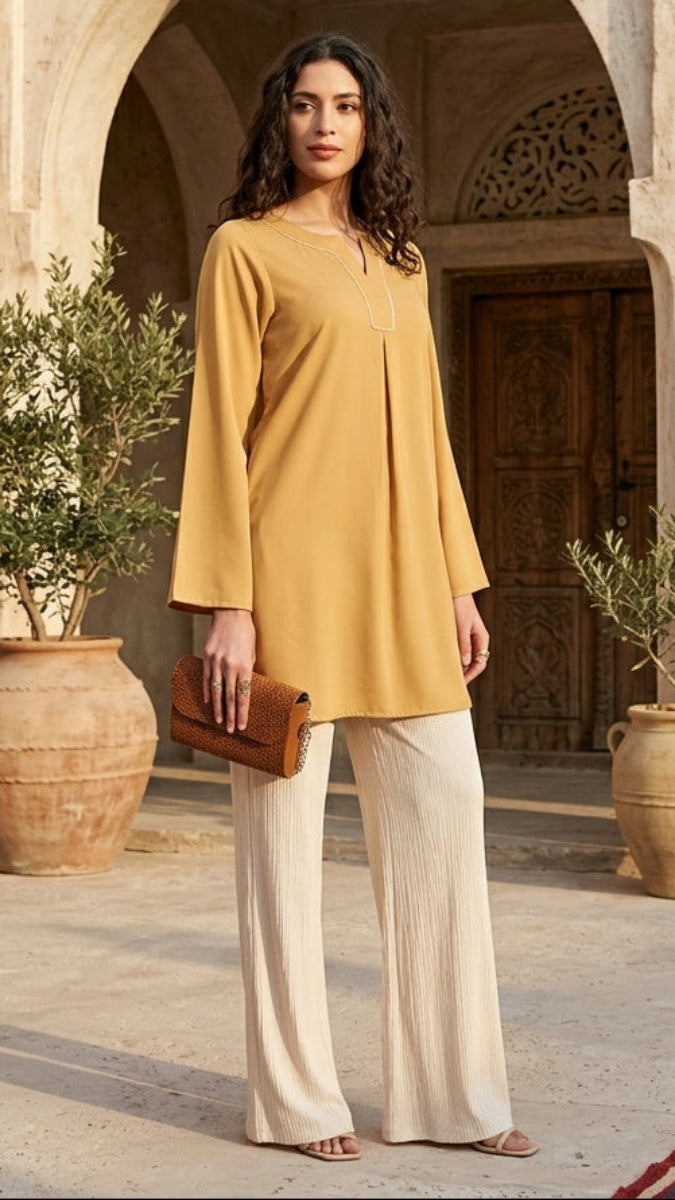 Woman in a mustard yellow top and beige pants standing in a sunlit courtyard with arches and potted plants.