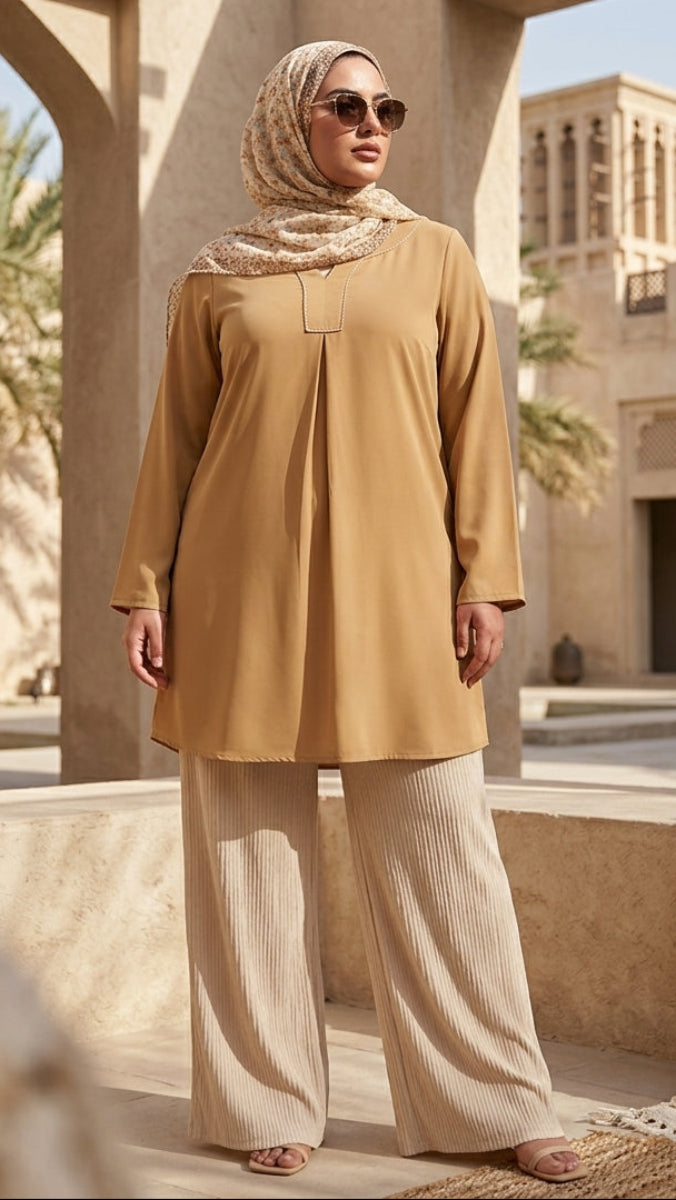 Woman in a mustard yellow top and beige pants standing in a sunlit courtyard with architectural elements.
