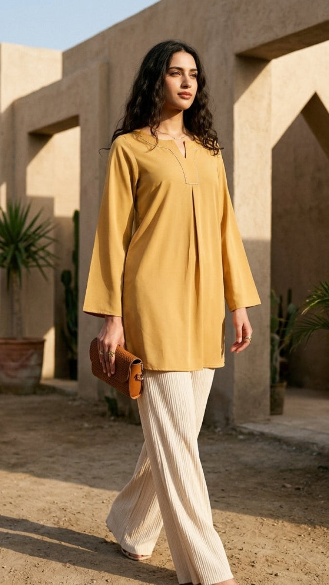 Woman in a yellow top and white pants walking outdoors with stone architecture and plants in the background