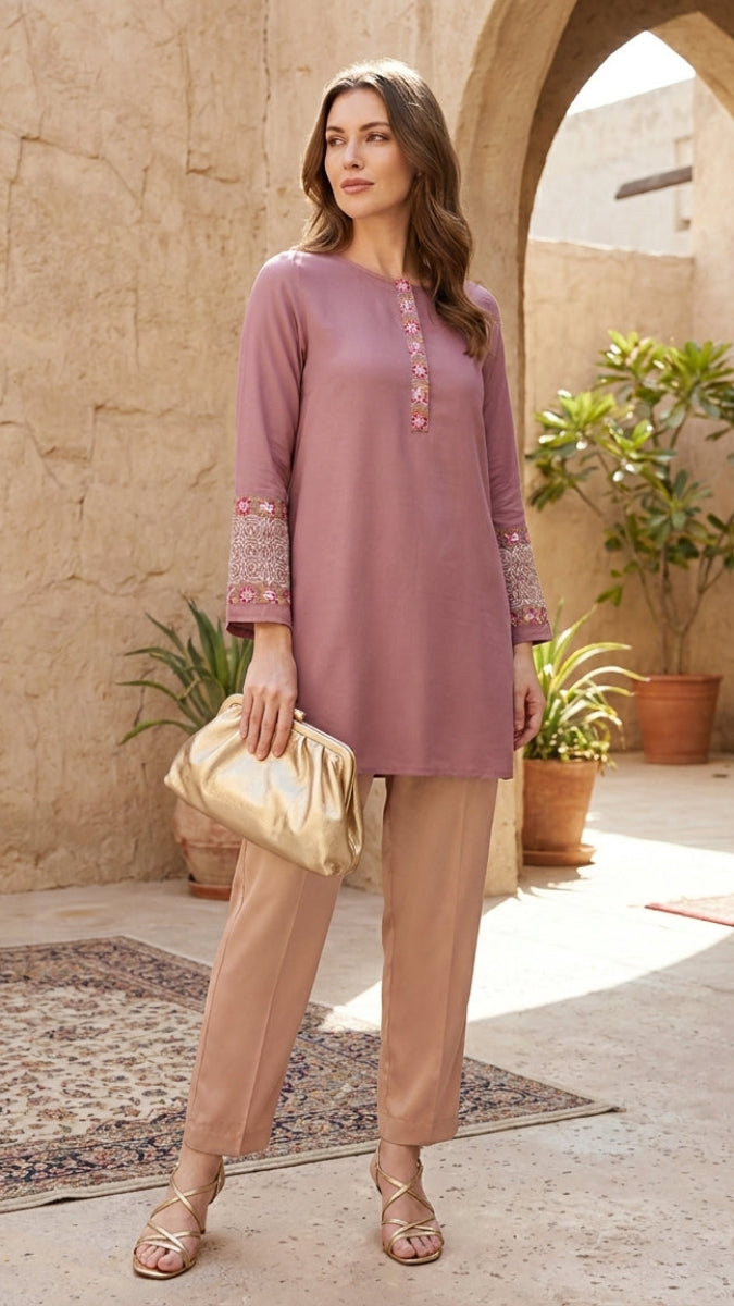 Woman in a pink top and beige pants standing in a sunlit courtyard with plants and arches.