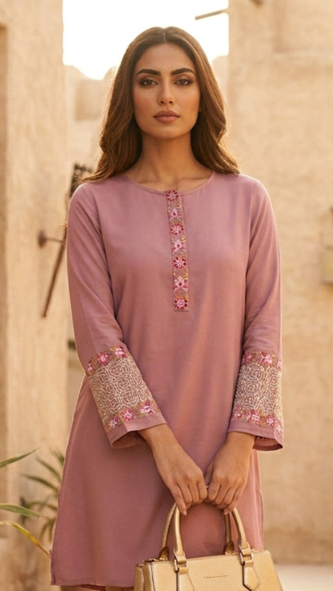 Woman in a pink dress holding a beige handbag in an outdoor setting with plants and stone walls.