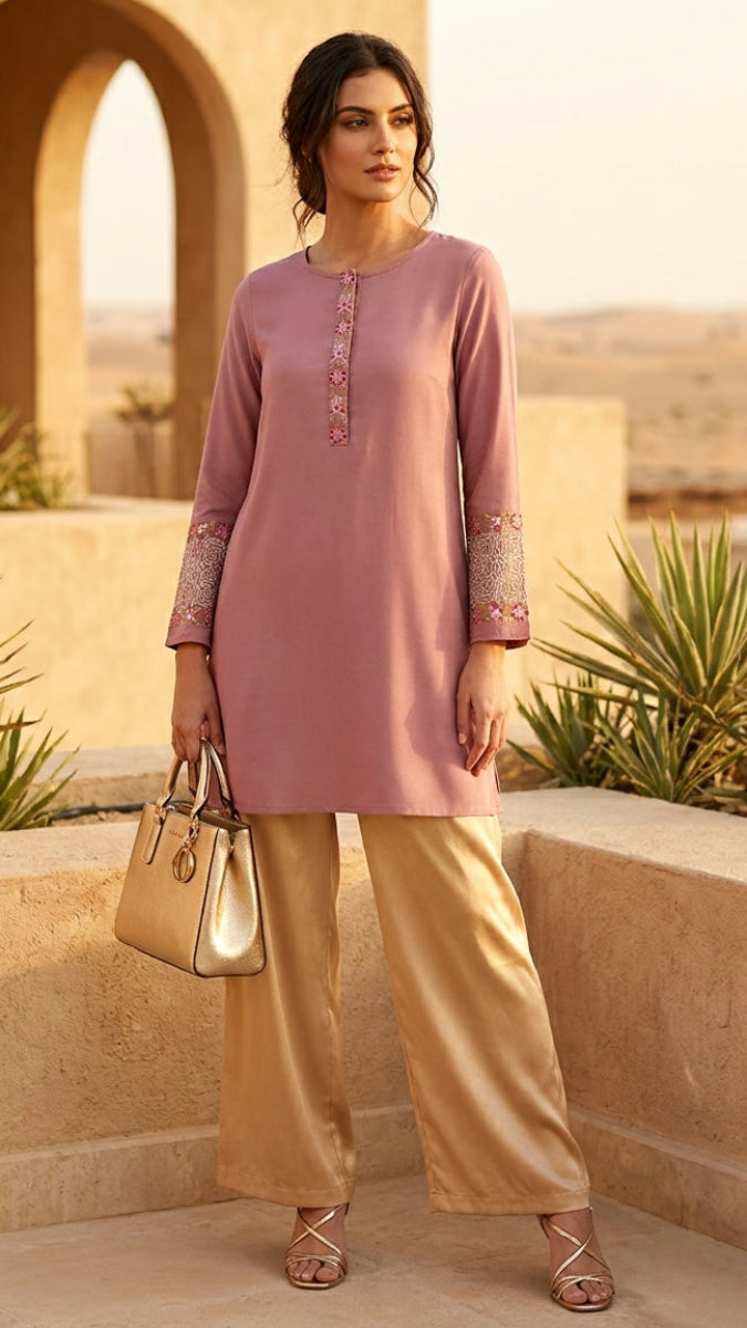 Woman in a pink top and beige pants standing in a desert setting with arches and plants.