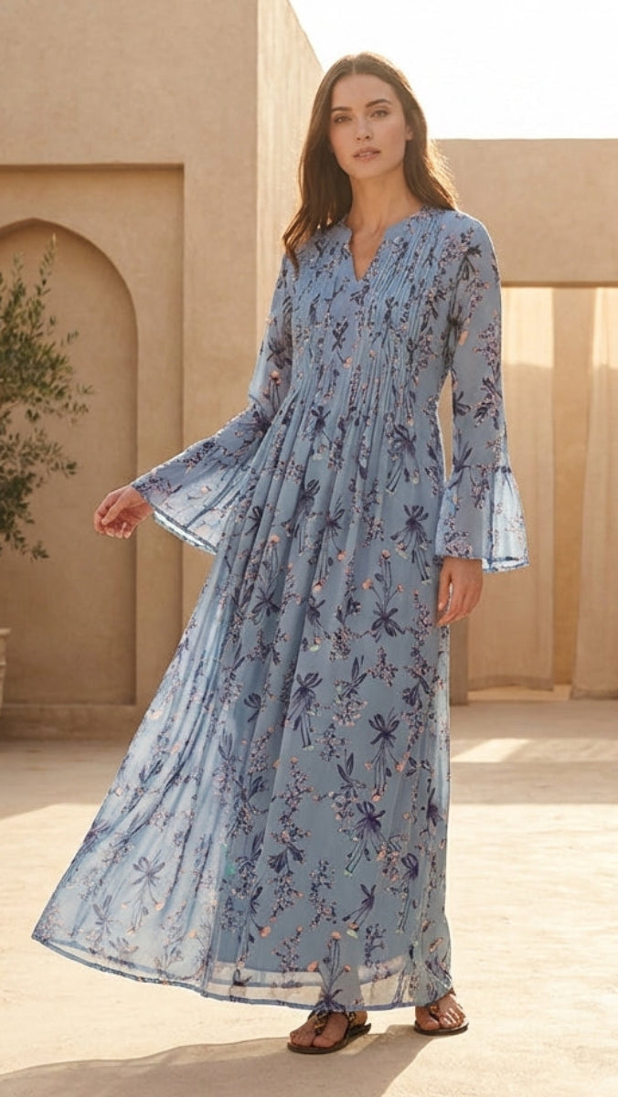 Woman in a blue floral dress standing in a sunlit courtyard with beige architecture and plants.