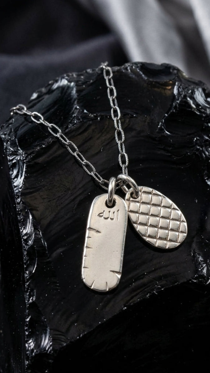 A silver pendant necklace displayed on a glossy black rock prop. The necklace features a fine link chain with two pendants: a flat rectangular bar engraved with '201' and a textured oval tag with a diamond-grid pattern.