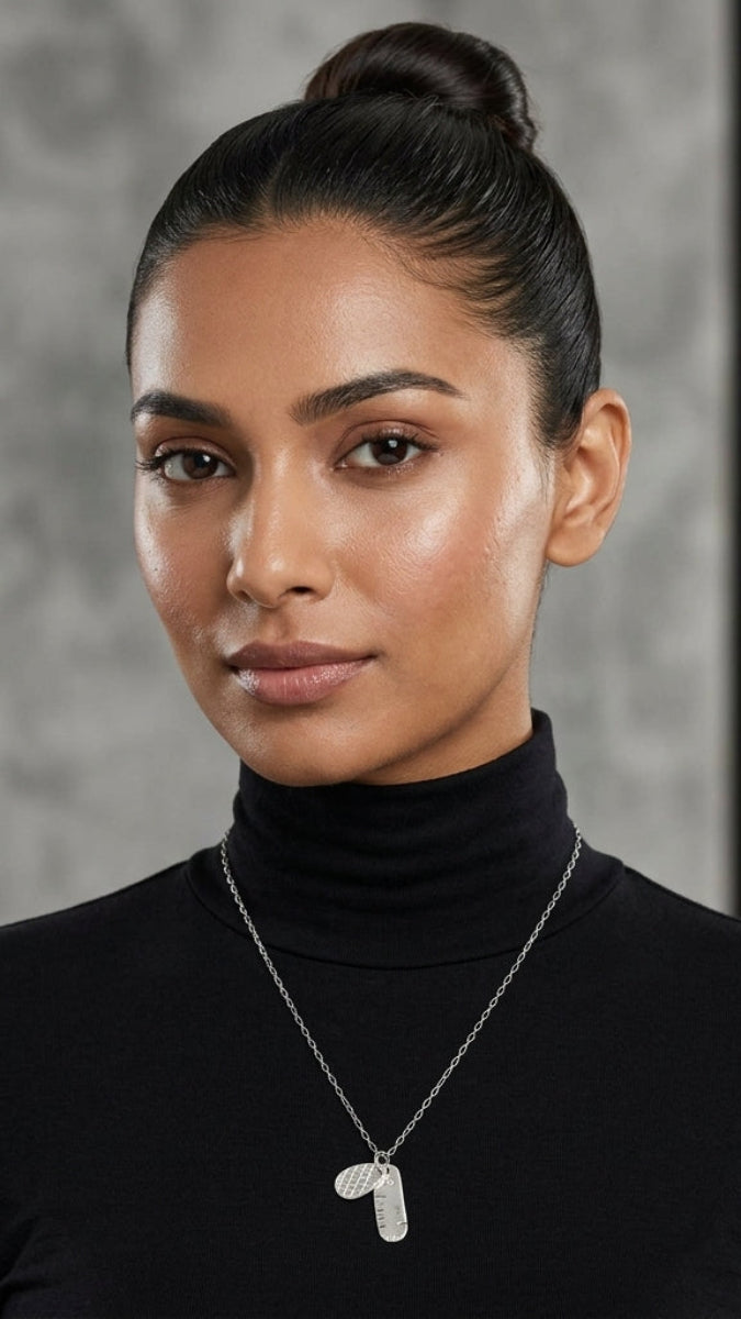 A woman with her hair in a bun wearing a black turtleneck and a silver pendant necklace with two small oval charms, photographed against a gray and black textured background.