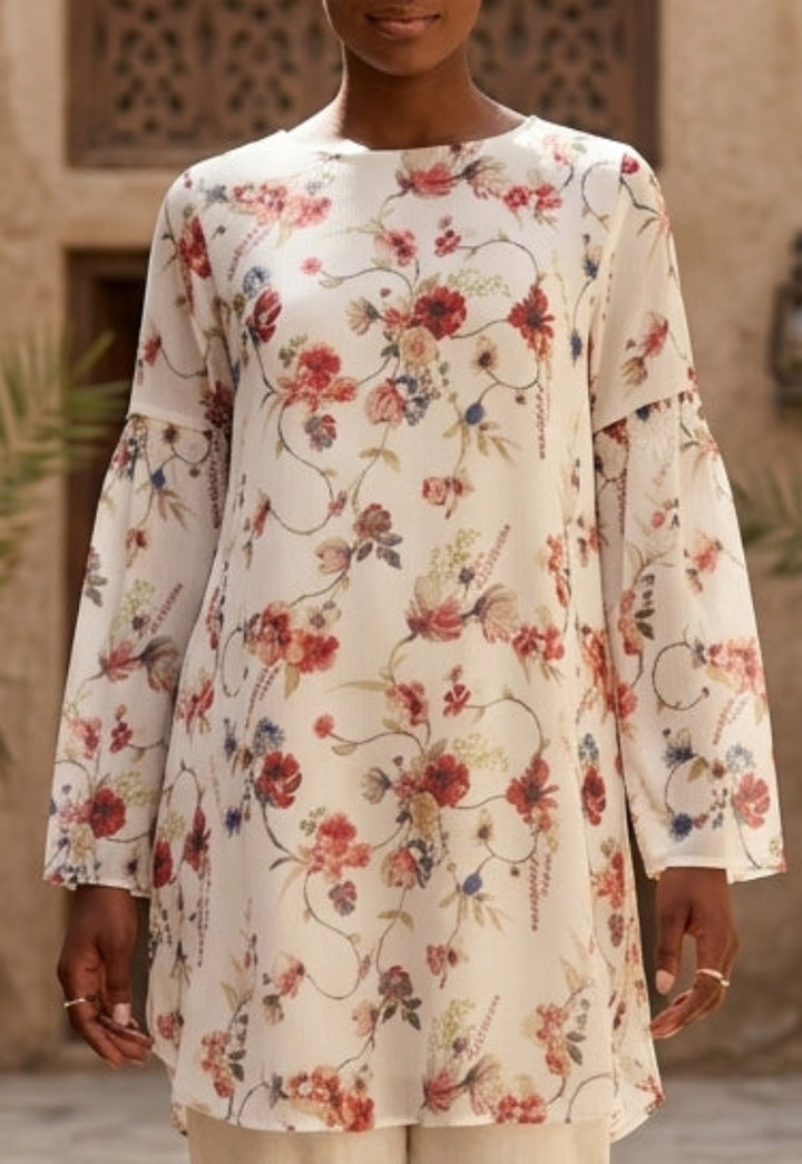 Woman in a floral top and beige pants walking in a sunlit courtyard.